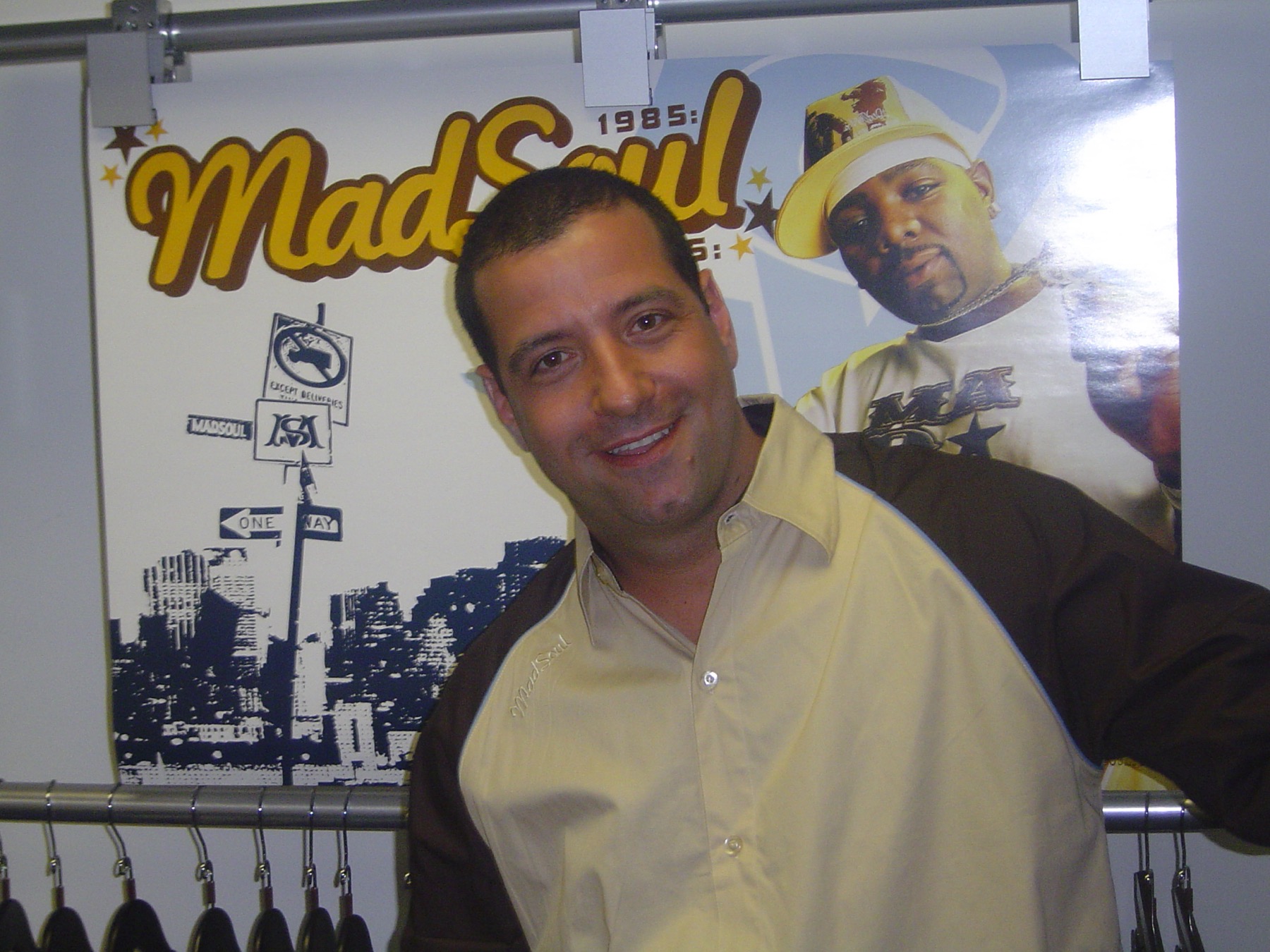 Marc D'Amelio smiling in the Madsoul showroom, standing in front of the brand's own 'MadSoul 4 ELMTS, Est. 1985' wall graphic, a rack of product beside him